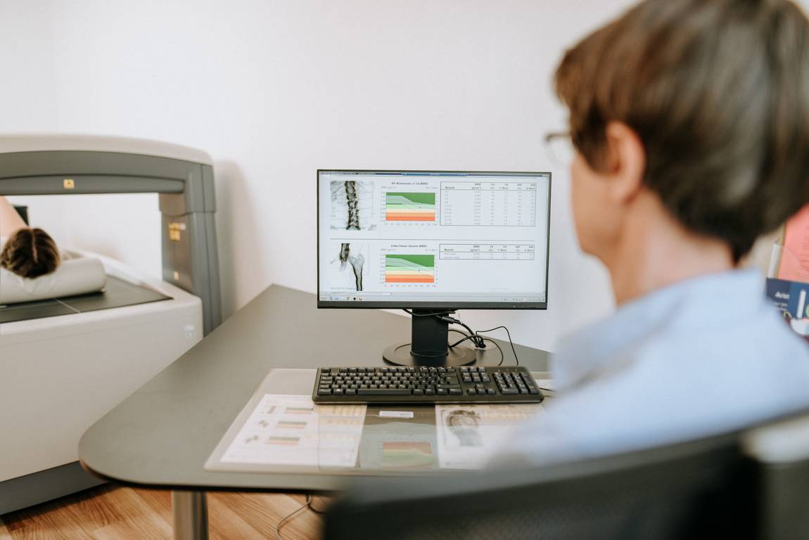 Ein Arzt prüft die Ergebnisse der Knochendichtemessung auf einem Computermonitor, während der Patient in einem medizinischen Untersuchungsraum auf einem Gerät liegt.