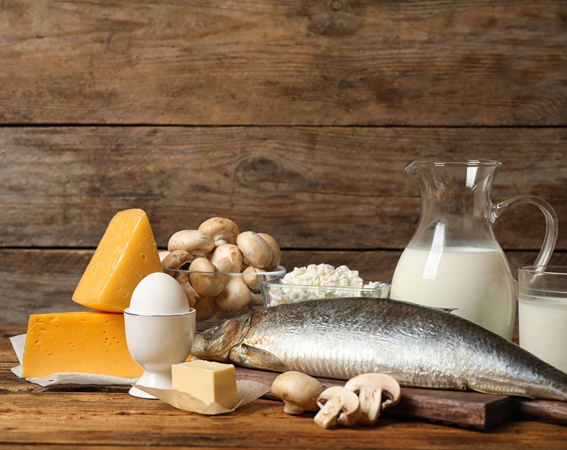 Eine Vielzahl von Vitamin-D-reichen Lebensmitteln auf einem Holztisch, darunter Käse, Pilze, Hüttenkäse, ein Glas und ein Krug Milch, ein ganzer Fisch, ein Ei in einem Eierbecher und ein Stück Butter.