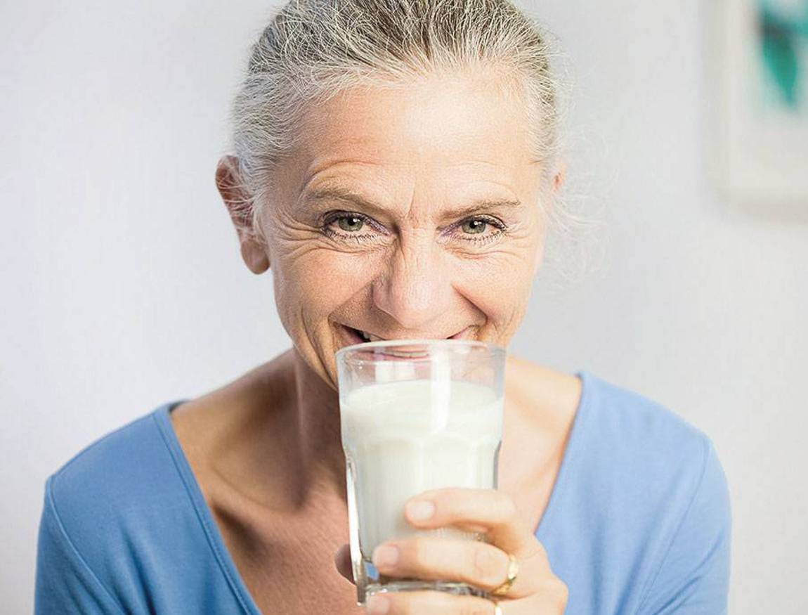 Eine ältere Frau mit grauem Haar lächelt, während sie ein Glas Milch an ihr Gesicht hält. Sie trägt ein hellblaues Hemd und befindet sich in einem Innenraum mit einem weichgezeichneten Hintergrund.